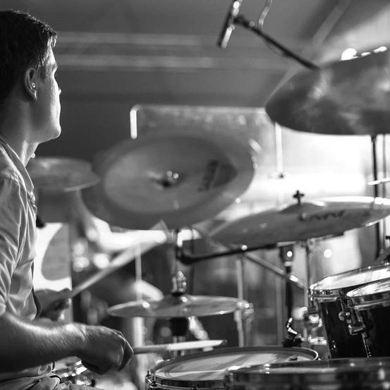 Ein Mann spielt Schlagzeug und konzentriert sich auf die Drumsticks in seinen Händen. Mehrere Becken sind hinter ihm aufgestellt, und ein Mikrofon ist darüber montiert. Die Umgebung scheint eine Aufführung oder ein Proben zu sein.