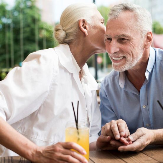 Ein älteres Paar sitzt an einem Tisch im Freien und tauscht einen liebevollen Kuss aus. Die Frau hält ein Glas mit Strohhalm und Orangensaft.