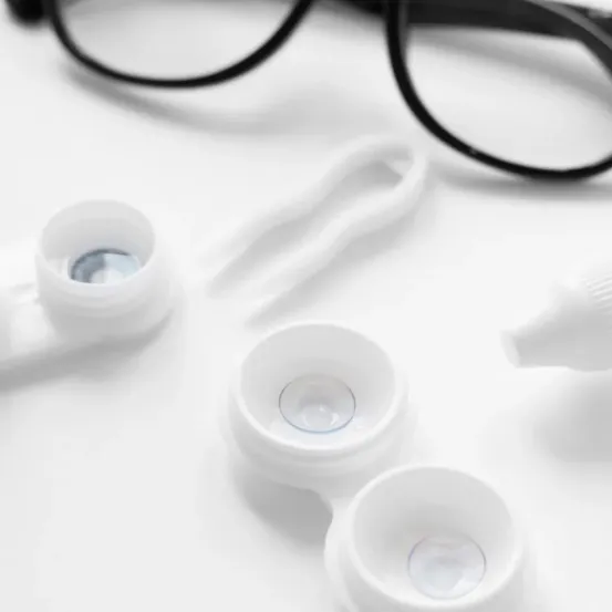 Ein Paar schwarze Brille, drei weiße Linsenbehälter und eine kleine Flasche Linsenlösung auf einer weißen Oberfläche.