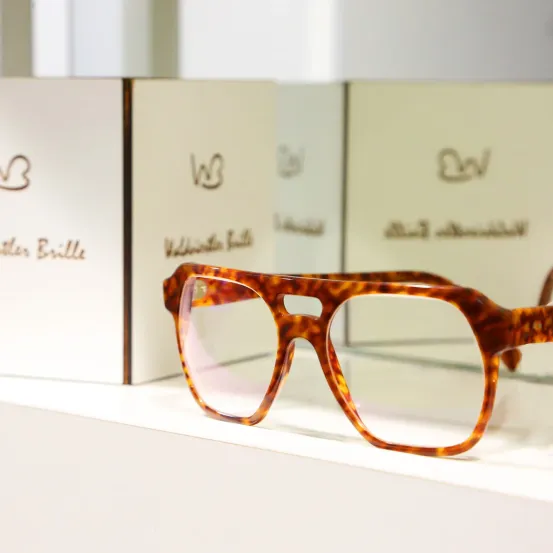 Ein Paar Schildkrötenbrille wird auf einem weißen Regal ausgestellt, mit vier weißen Schachteln dahinter, jede mit einem einzigartigen Design und Text versehen.