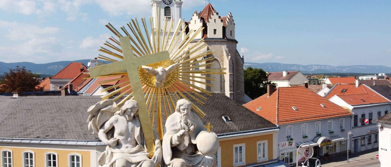 Ein goldenes Kreuz mit einer Taube darüber, umgeben von Strahlen, steht vor einer Statue zweier Figuren. Dahinter befindet sich eine Kirche mit einem Glockenturm und roten Dächern.