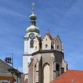 Eine historische Kirche mit einem hohen Turm und einer Uhr, in einer Stadt gelegen, mit einem rotgedeckten Gebäude in der Nähe.