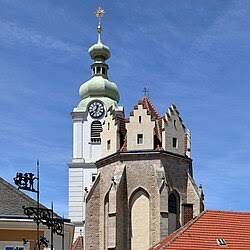 Eine historische Kirche mit einem hohen Turm und einer Uhr, in einer Stadt gelegen, mit einem rotgedeckten Gebäude in der Nähe.