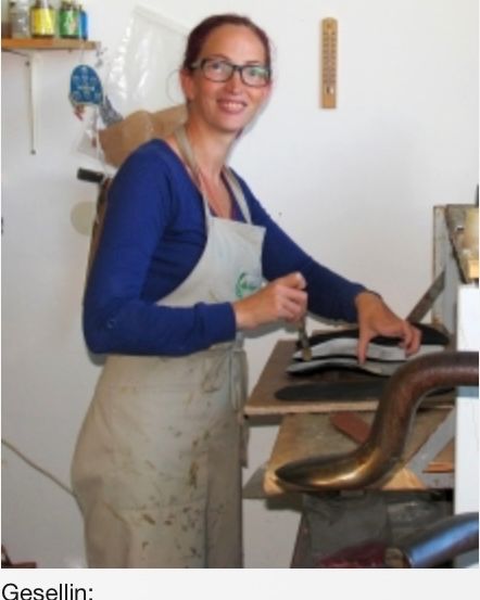 Eine Frau in einem weißen Schürze steht in einer Werkstatt und hält ein Werkzeug in der Hand. Hinter ihr befindet sich ein Tisch mit Schuhen und einer Maschine.