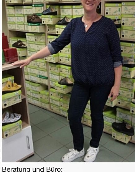 Eine Frau steht in einem Schuhgeschäft, trägt eine blaue Bluse und schwarze Hose. Hinter ihr befinden sich mit Schuhkartons gefüllte Regale, und sie lächelt und posiert für ein Foto.