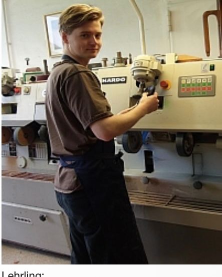 Ein Mann in einer Werkstatt bedient eine Maschine mit der Aufschrift 'HARDO', während er ein braunes Hemd und eine Schürze trägt. Hinter ihm ist eine Maschine mit verschiedenen Rädern und einem Bedienfeld zu sehen, zusammen mit einem gerahmten Bild an der Wand.