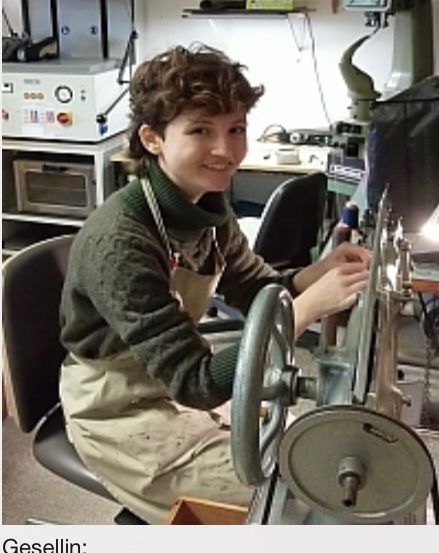 Eine junge Frau mit lockigem Haar, die einen grünen Pullover und eine Schürze trägt, arbeitet an einer Maschine in einer Werkstatt. Die Maschine ist groß mit einem runden Griff. Hinter ihr steht ein Schreibtisch mit verschiedenen Werkzeugen und einer Maschine darauf.