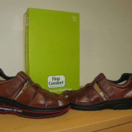 Ein Paar braune Ledersandalen mit schwarzer Sohle und rotem Streifen liegen auf einer Holzoberfläche neben einer grünen Box mit der Aufschrift 'Finn Comfort'.