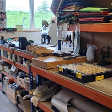 Ein Werkstattraum gefüllt mit Werkzeugen und Materialien, einschließlich eines Regals mit verschiedenen Gegenständen wie Schuhen, einem Werkzeugkasten und einer Aufbewahrungsbox. Der Raum hat ein Glasfenster mit Blick auf ein Grasfeld.