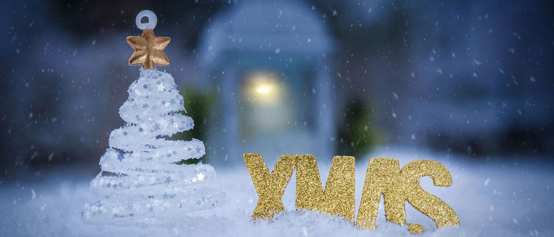 Ein kleiner glitzernder Weihnachtsbaum mit einem goldenen Stern darauf steht im Schnee. Daneben ist das Wort 'XMAS' in Goldglitter geschrieben. Im Hintergrund leuchtet ein Laternenlicht schwach.