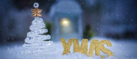 Ein kleiner glitzernder Weihnachtsbaum mit einem goldenen Stern darauf steht im Schnee. Daneben ist das Wort 'XMAS' in Goldglitter geschrieben. Im Hintergrund leuchtet ein Laternenlicht schwach.