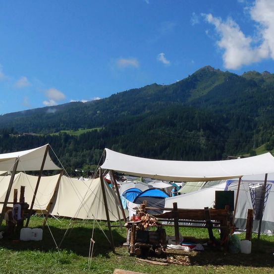 Ein Campingplatz mit mehreren aufgestellten Zelten auf Grasboden, mit einem Holztisch und Feuerholz. Berge mit blauem Himmel und Wolken im Hintergrund.