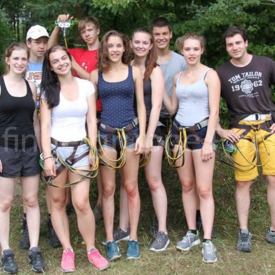 Eine Gruppe junger Menschen, sowohl männlich als auch weiblich, posiert für ein Foto, während sie Gurtzeuge und Turnschuhe tragen. Sie scheinen sich in einem Waldgebiet zu befinden.