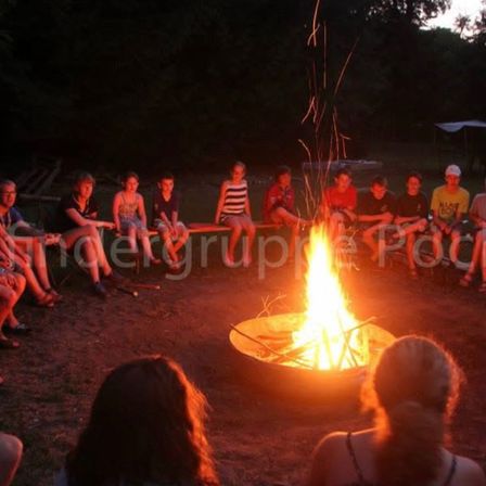 Eine Gruppe von Menschen sitzt bei Nacht um ein Lagerfeuer, mit einem Zelt im Hintergrund. Einige tragen Hüte, und sie werden vom Feuerschein beleuchtet.