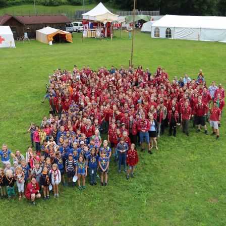 Eine große Gruppe von Menschen in roten und blauen Uniformen posiert für ein Foto auf einem Rasenfeld. Im Hintergrund befinden sich Zelte und ein rotes Kreuzzelt.