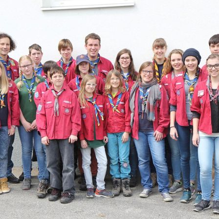 Eine Gruppe junger Pfadfinder in roten Jacken steht vor einer weißen Wand und lächelt für ein Foto. Sie sind in Alter und Geschlecht unterschiedlich, einige tragen Brillen und Mützen.