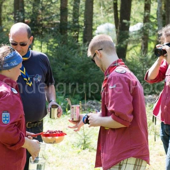 Eine Gruppe von Menschen in roten Hemden versammelt sich im Wald. Eine Frau hält eine Schüssel mit roten Beeren und ein Mann eine Dose. Ein anderer Mann macht ein Foto.