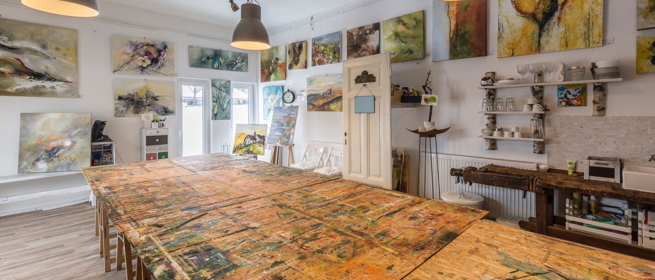Ein Studio mit einem langen Holztisch, der mit Farbe bedeckt ist, verschiedenen Gemälden an den Wänden und Lampen, die von der Decke hängen.