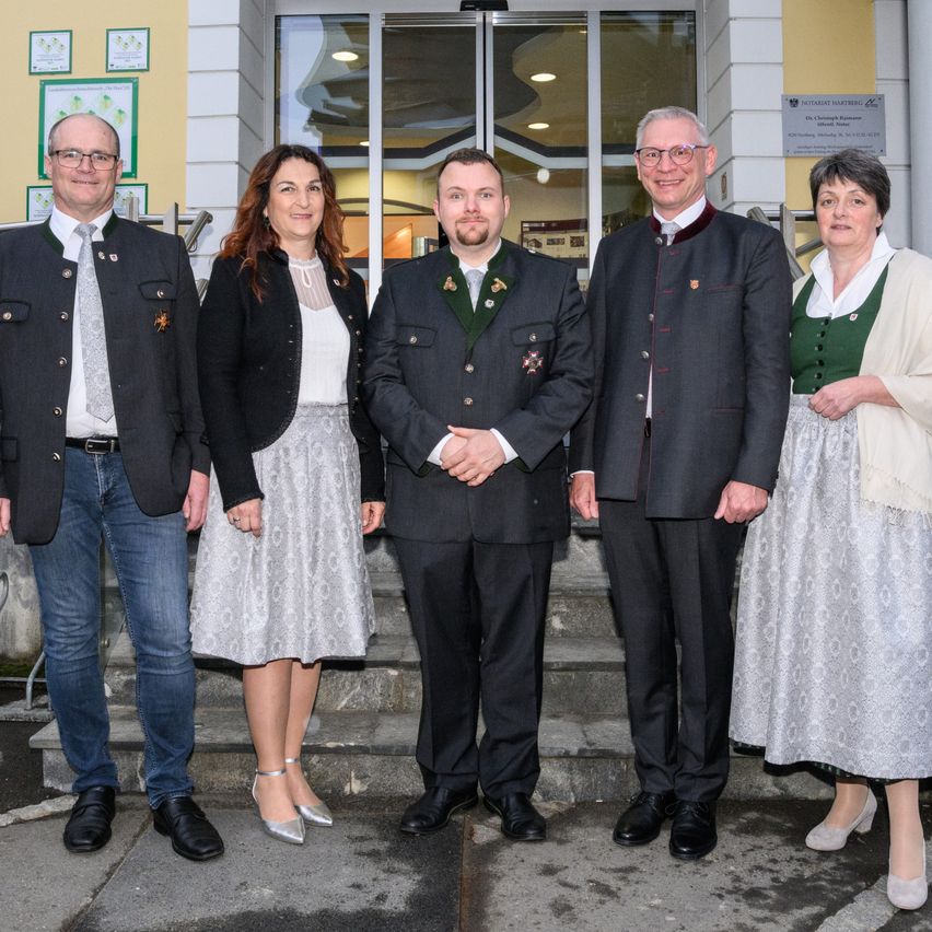 Fünf Personen in traditioneller Kleidung stehen vor einem Gebäude mit Glastüren. Zwei Männer tragen Anzüge mit Krawatten und Medaillen, während die anderen Kleider tragen.