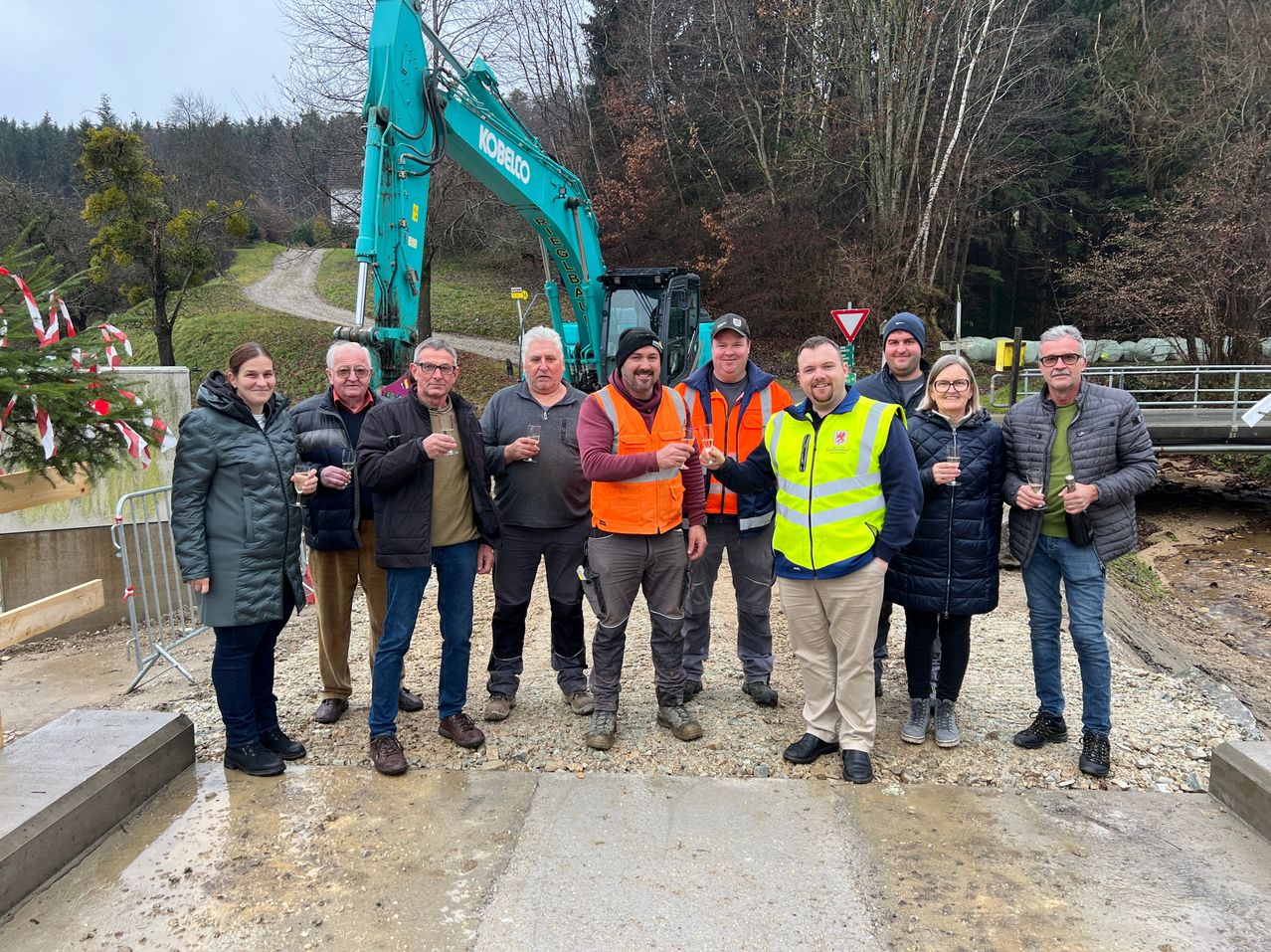 Eine Gruppe von Menschen in Winterkleidung steht vor einem grünen Baufahrzeug. Sie halten alle Gläser mit Wein und scheinen ein Ereignis zu feiern. Im Hintergrund ist eine Feldstraße und Bäume zu sehen.