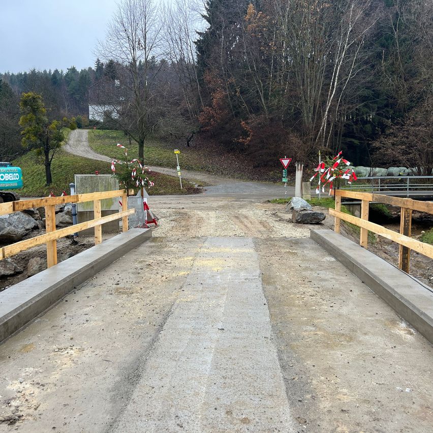 Eine Baustelle zeigt eine neue Brücke im Bau. Holzzäune umranden die Brücke, und Warnschilder sind angebracht. Die Straße ist teilweise asphaltiert.