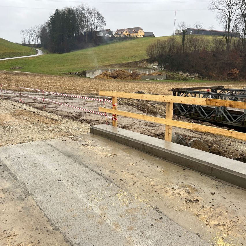 Eine Baustelle zeigt eine neue Betonstraße mit einer hölzernen Barriere. Dahinter ist eine hügelige Landschaft mit einem gelben Haus und einer Straße zu sehen. Bäume und Büsche umgeben das Gebiet.