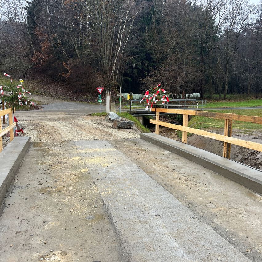 Eine unbefestigte Straße mit einer neu errichteten Betonbrücke über einem kleinen Bach. Die Brücke hat hölzerne Geländer auf beiden Seiten. In der Nähe stehen Bäume, und eine rot-weiße Flagge ist an einem Pfahl befestigt.