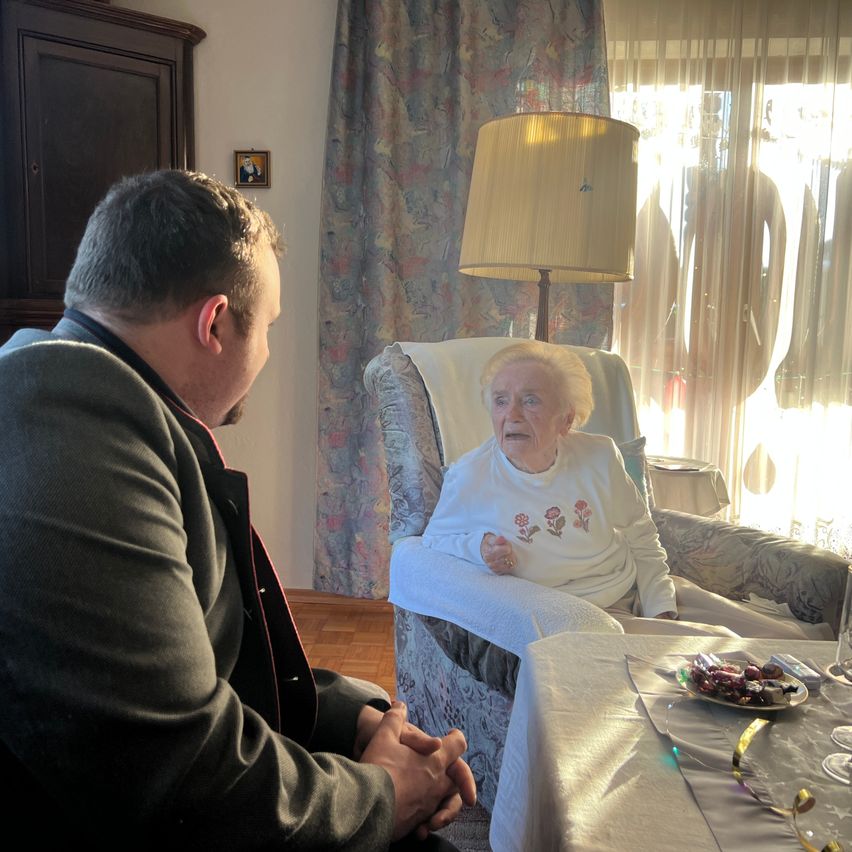 Eine ältere Frau sitzt auf einem gepolsterten Stuhl mit lächelndem Gesicht, ein Mann sitzt ihr gegenüber. Ein Tisch mit einem Teller mit Süßigkeiten steht vor ihnen. Eine Lampe ist auf dem Boden platziert.