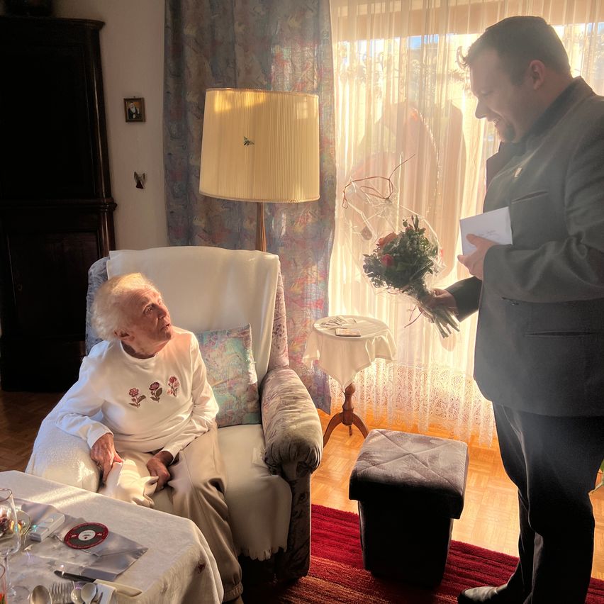 Eine ältere Frau sitzt auf einem Stuhl und lächelt, während ein Mann ihr einen Blumenstrauß überreicht. Die Szene spielt sich drinnen ab, mit einer Lampe und einem Fenster im Hintergrund.