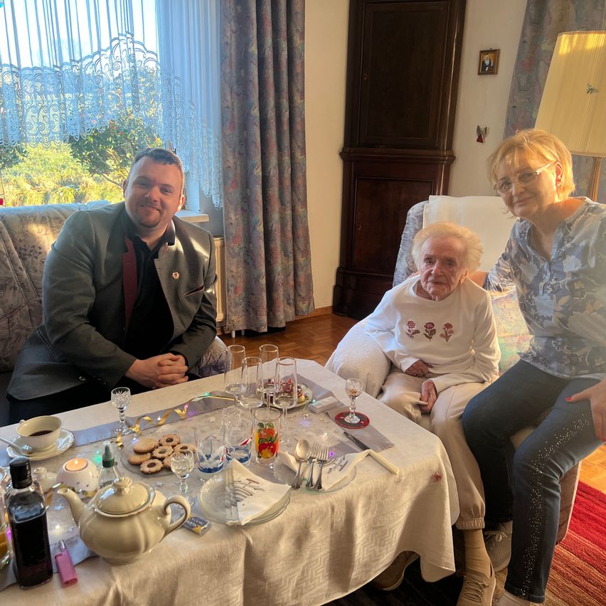 Drei Menschen sitzen in einem Wohnzimmer. Eine Frau sitzt auf einem Stuhl und eine andere Frau auf einer Couch. Sie lächeln. Vor ihnen steht ein Tisch mit einem Teepott, Weingläsern und anderen Gegenständen. Eine Lampe befindet sich auf der rechten Seite des Raumes.