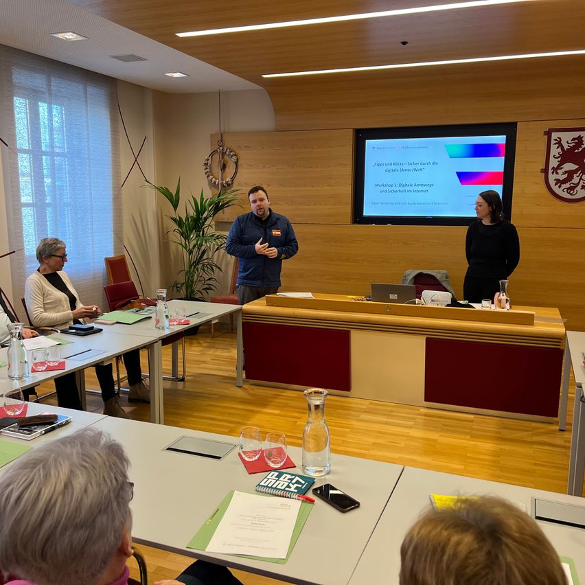 Ein Raum für Besprechungen mit Holzboden. Ein Mann spricht, während eine Frau am Podium steht. Hinter ihnen befindet sich ein Präsentationsbildschirm. Teilnehmer sitzen an Tischen.