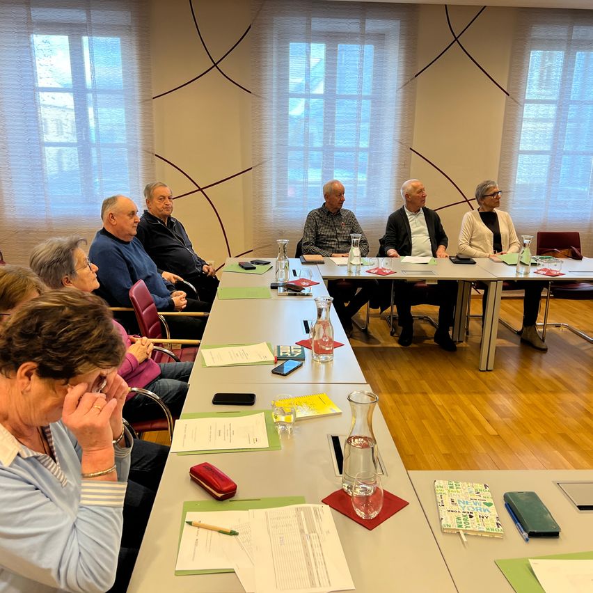 Eine Gruppe älterer Personen sitzt um einen Tisch in einem Konferenzraum, mit Papieren, Stiften und Gläsern auf dem Tisch. Der Raum hat große Fenster mit Vorhängen und einen Holzboden.