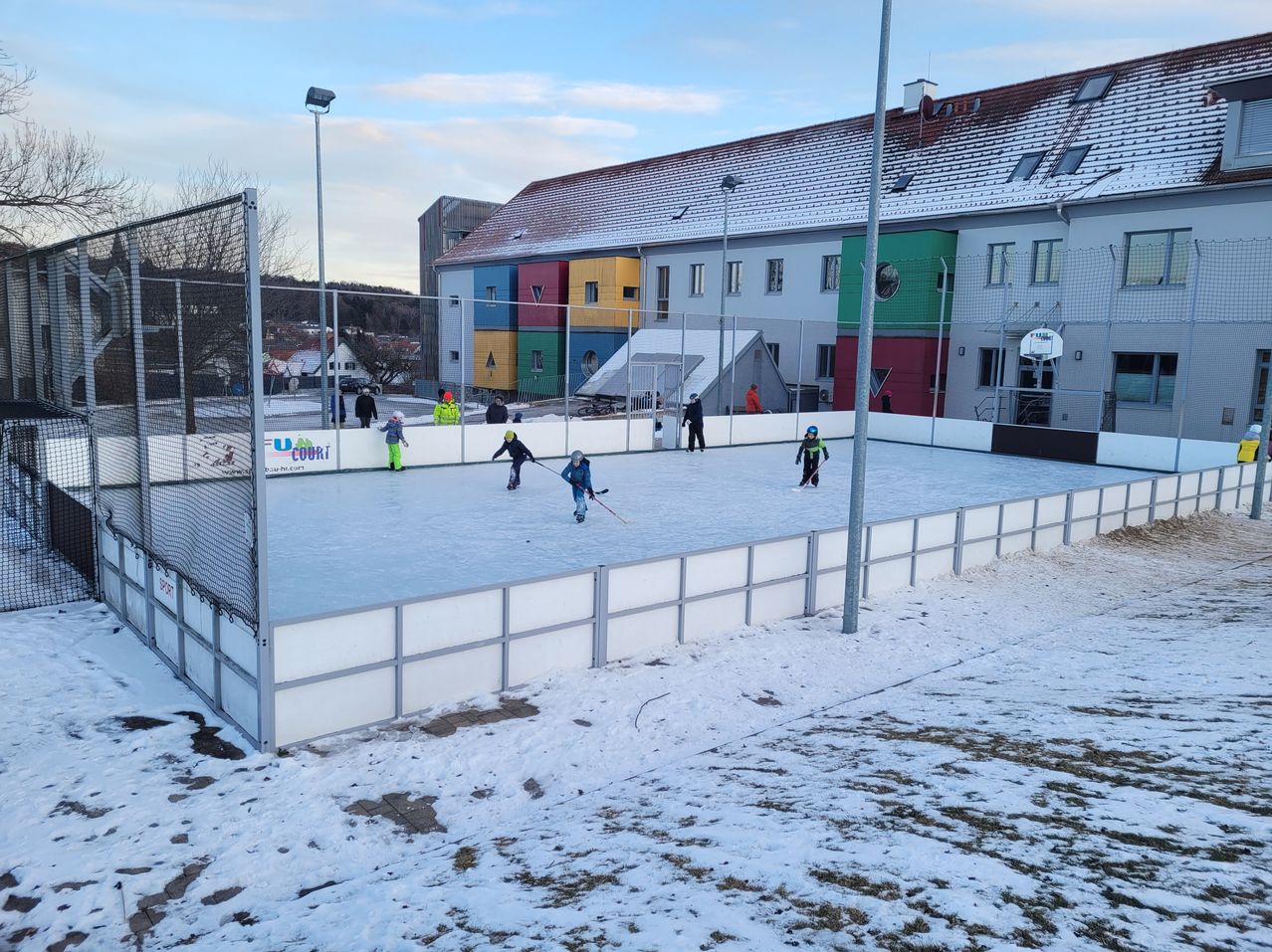 Mehrere Personen spielen Eishockey auf einer Outdoor-Eisbahn, die von einem Zaun umgeben ist, bei schneereichem Wetter. Hinter dem Zaun befindet sich ein Gebäude mit farbenfrohen Wänden.