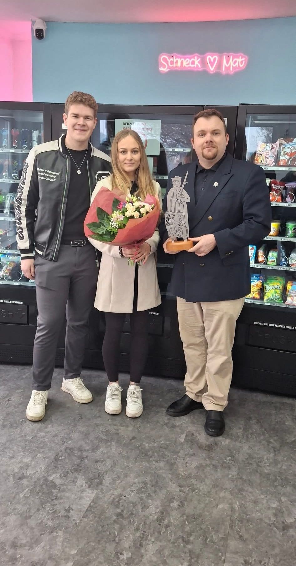 Drei Personen stehen vor einem Verkaufsautomaten, der Mann auf der rechten Seite hält eine Trophäe, die Frau einen Blumenstrauß und der Mann auf der linken Seite trägt eine Halskette.