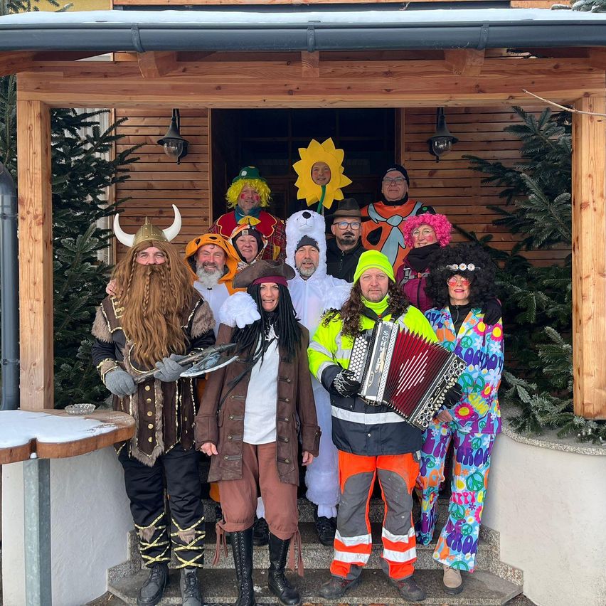 A group of people dressed in various costumes, including a Viking, a pirate, a bear, and a sun, stand in front of a wooden house. One person plays an accordion.