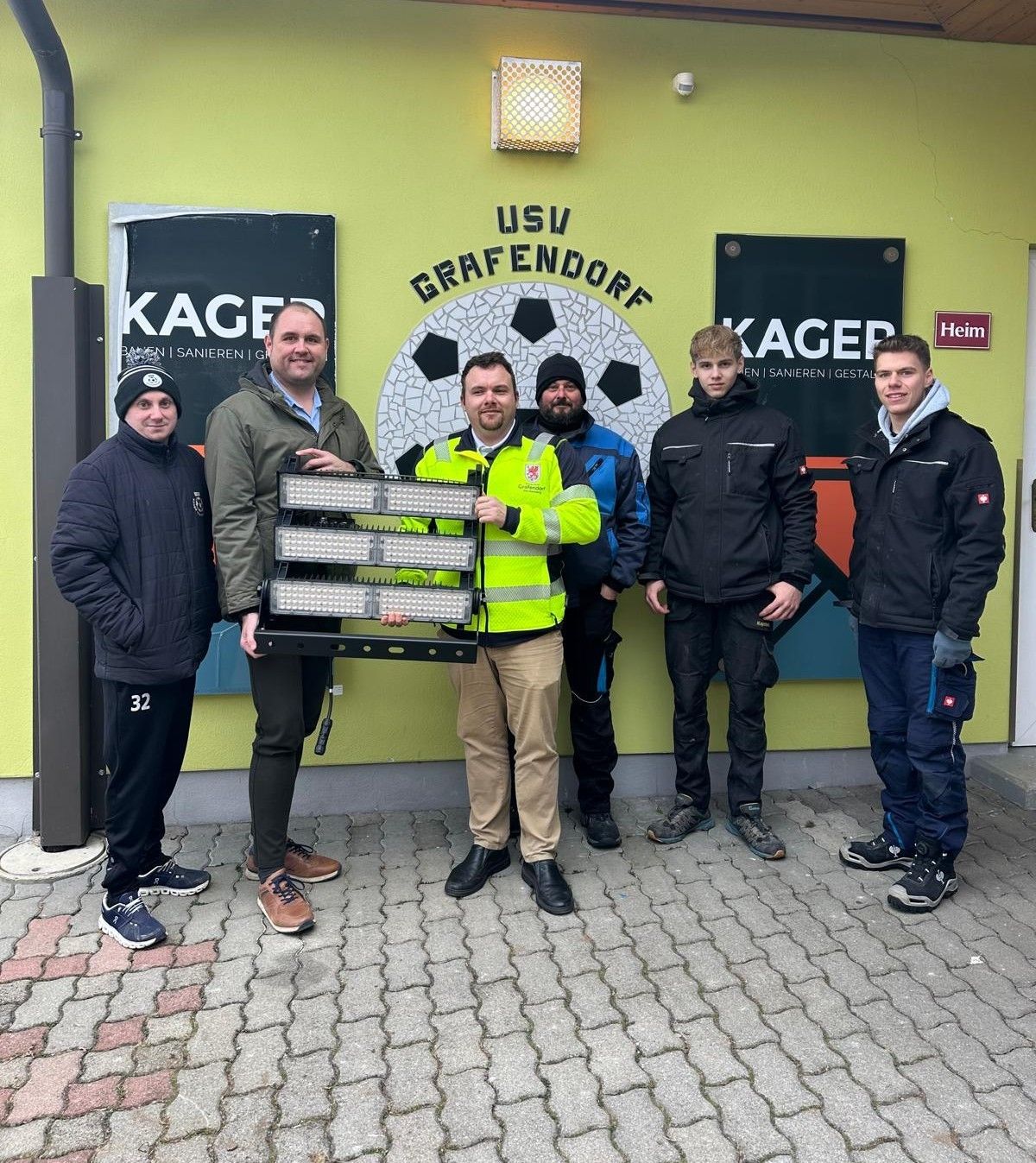 Sechs Männer stehen vor einem Gebäude mit gelben Wänden und schwarzen Bannern. Einer hält eine Lampe. Sie tragen Winterkleidung.