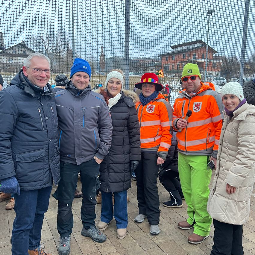 Sechs Menschen in Winterkleidung stehen draußen. Ein Mann mit Brille lächelt. Ein Mann in einem blauen Hut hält ein Mikrofon. Dahinter ist ein Netz und Gebäude.