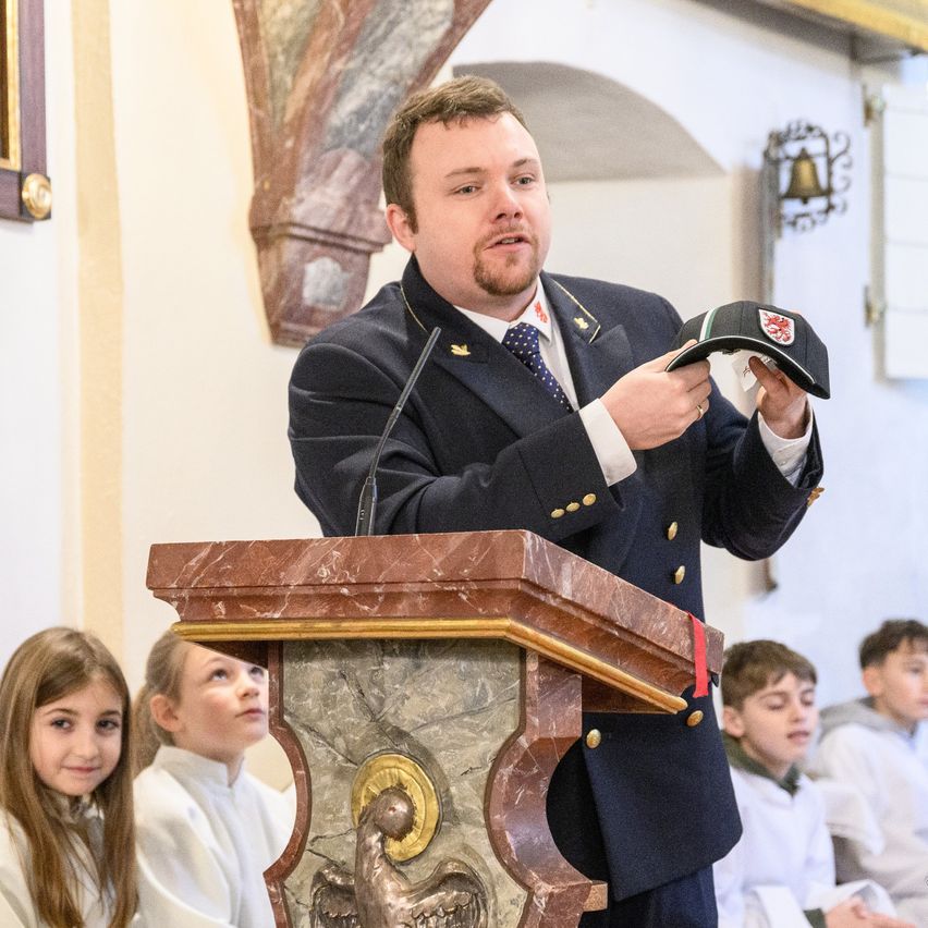 Ein Mann in einer Marineuniform spricht an einem Podium, während er einen Hut hält. Mehrere Kinder hören aufmerksam zu.