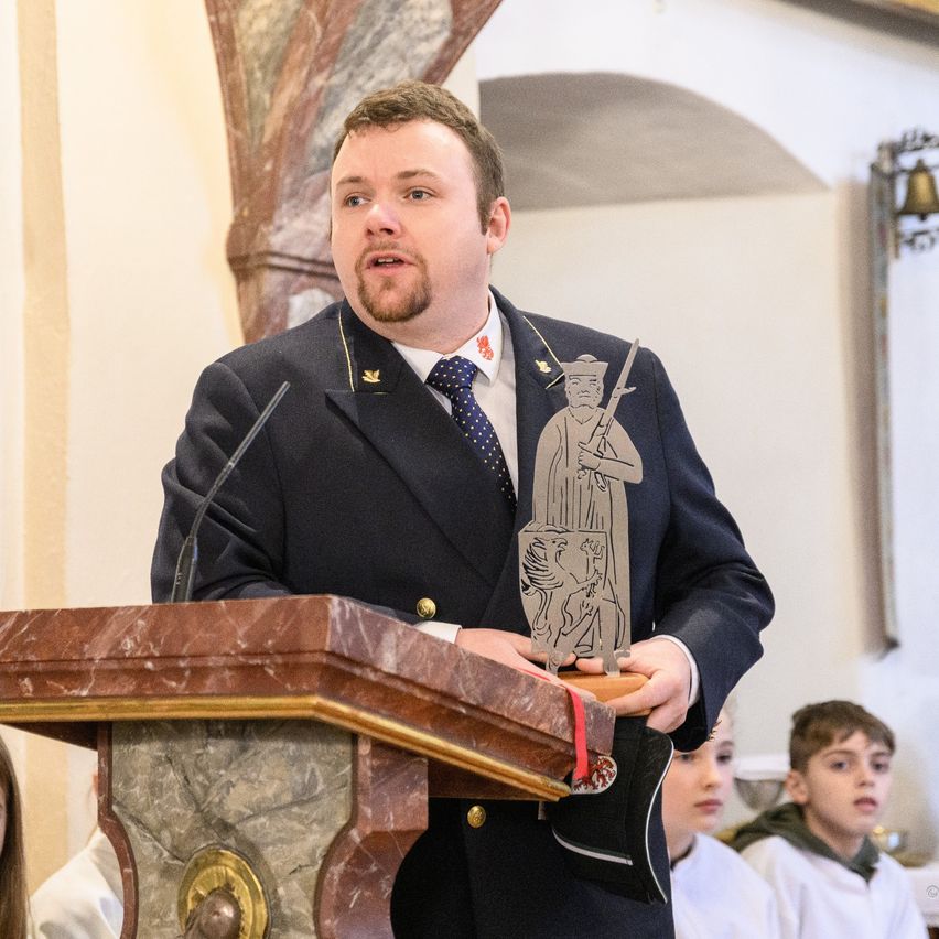 Ein Mann in einem Anzug spricht an einem Podium und hält eine Trophäe mit einer Figur darauf. Dahinter stehen zwei Kinder und schauen sich etwas an.