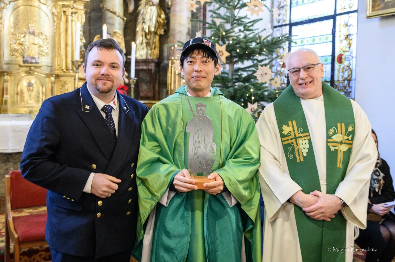 Drei Männer stehen zusammen vor einem geschmückten Weihnachtsbaum. Der Mann auf der linken Seite trägt einen dunkelblauen Anzug mit goldenen Knöpfen, und der Mann in der Mitte hält einen Pokal. Der Mann auf der rechten Seite trägt einen grünen und weißen Umhang.