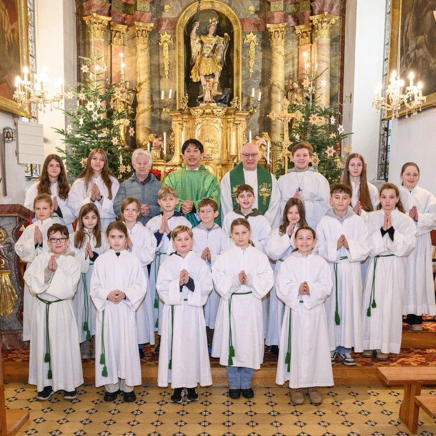 Eine Gruppe von Kindern und Erwachsenen in weißen Gewändern steht in einer Kirche, betet und schaut auf den Altar mit einer goldenen Statue einer Frau.