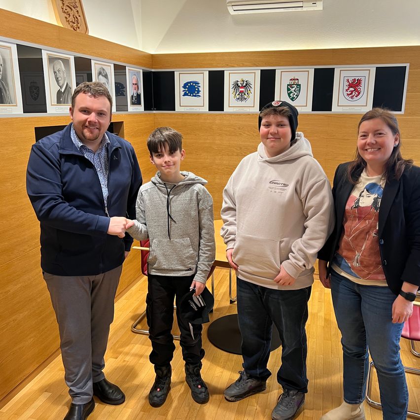 Vier Menschen, drei Erwachsene und ein Junge, stehen in einem Raum. Die Erwachsenen lächeln, und einer von ihnen schüttelt dem Jungen die Hand. Hinter ihnen hängen gerahmte Bilder und Logos an der Wand.