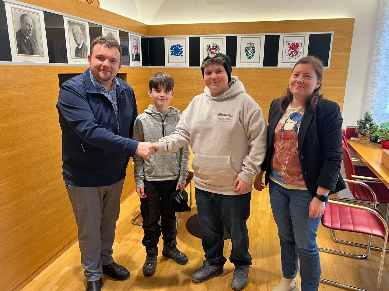 Vier Personen, zwei Männer und zwei Frauen, stehen in einem Raum und lächeln. Sie scheinen für ein Foto zu posieren. Der Mann auf der linken Seite schüttelt einem anderen Mann die Hand. Hinter ihnen sind mehrere Bilderrahmen an der Wand montiert. Ein roter Stuhl steht auf der rechten Seite.