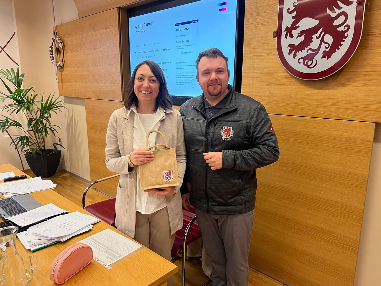 Eine Frau und ein Mann stehen in einem Büroraum mit einem Bildschirm, auf dem 'App ID Austria' angezeigt wird. Die Frau hält eine Tasche und lächelt, während der Mann mit der Hand gestikuliert.