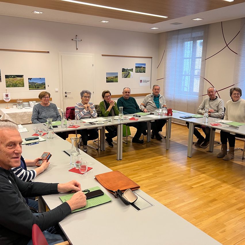 Eine Gruppe von Menschen sitzt in einem Konferenzraum, möglicherweise in einer Besprechung. Der Raum hat einen Holzboden, eine weiße Wand und ein Kreuz an der Wand. Die Menschen sitzen vor einem langen Tisch mit Gläsern, Flaschen und anderen Gegenständen. Die Frau links hält ein Buch, und der Mann rechts trägt einen Rucksack.