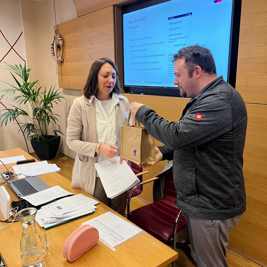 Ein erwachsener Mann und eine Frau in einer Büroumgebung. Der Mann reicht der Frau einen braunen Papierbeutel. Der Schreibtisch hat einen Laptop, eine Karaffe und Papiere. Eine Pflanze steht in der Ecke.