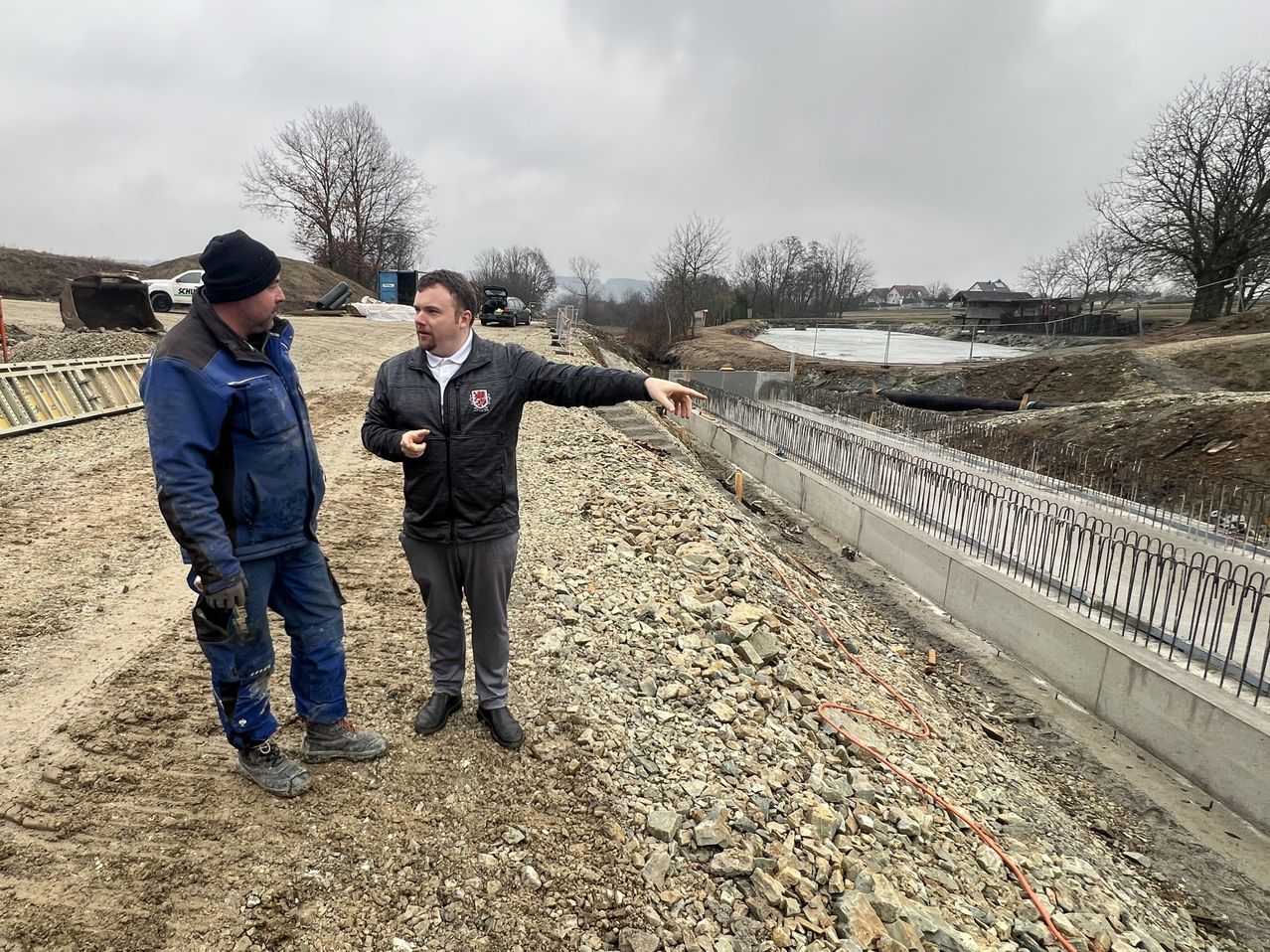 Zwei Männer stehen auf einem Schotterbereich mit einer Betonbarriere und zeigen auf das Gelände. Dahinter befindet sich ein Gewässer mit Bäumen und Häusern.