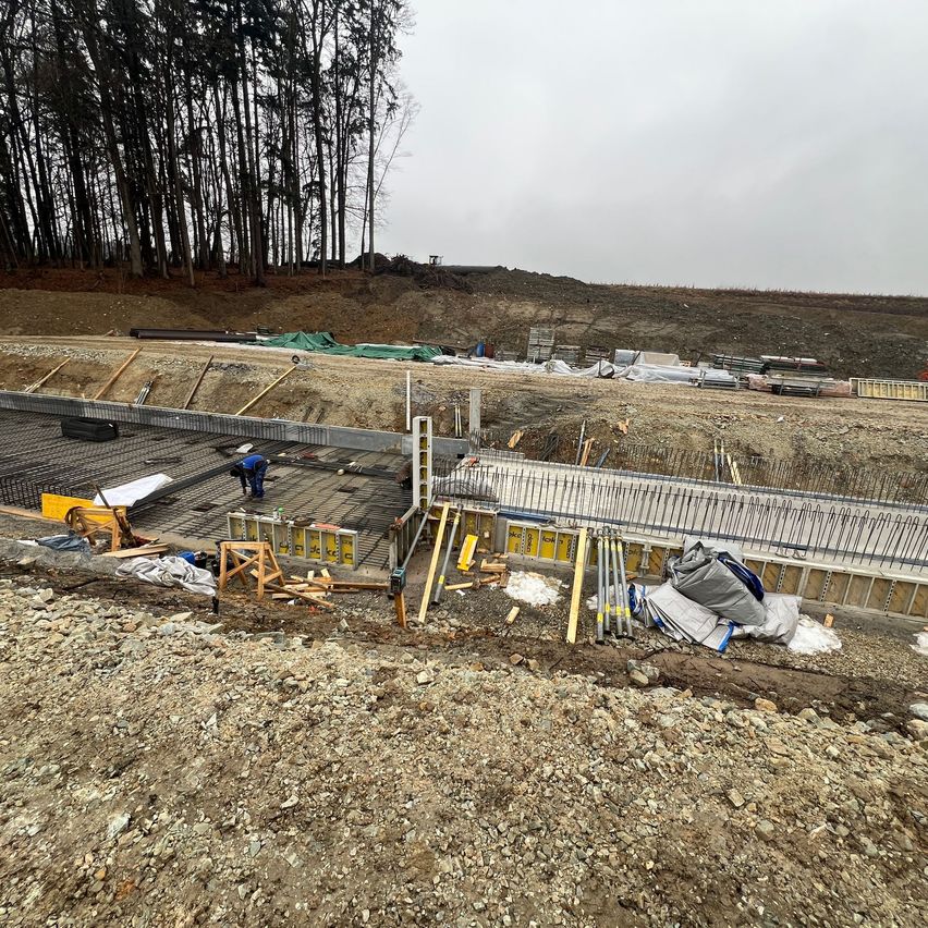 Eine Baustelle mit Arbeitern, einem Fundament und Materialien. Im Hintergrund befinden sich Bäume und eine Straße.