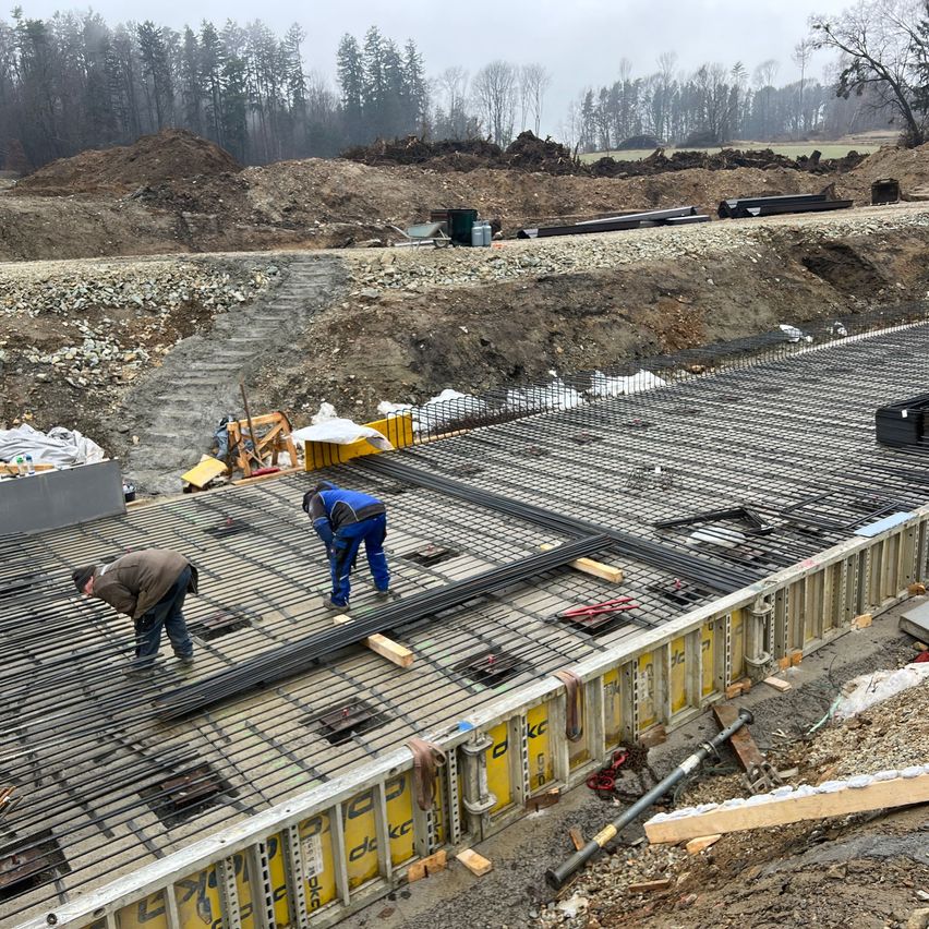 Zwei Arbeiter bauen eine Betonfundierung an einer Baustelle. Stahlstäbe liegen auf dem Boden. Schmutz und Steine sind verstreut.