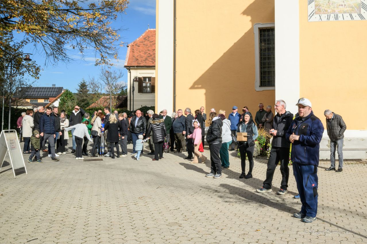 A group of people, including adults and children, stand in line in front of a building with a red roof and a yellow wall.